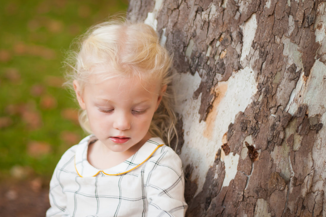 Little girl looks down sweetly while reclining against a tree