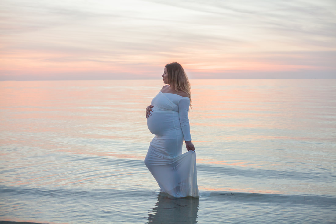 maternity photo of woman wearing white gown stands in the gulf of mexico with a pastel sunset behind her