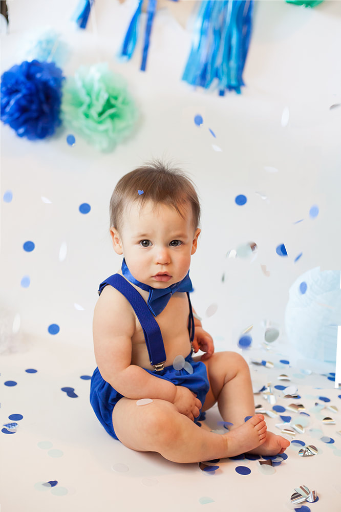 Baby boy in blue suspenders looks into the camera while blue and white confetti falls all around him