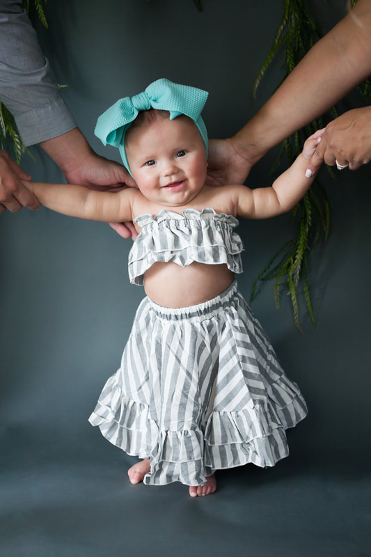 Smiling baby girl standing and holding onto her parents hands wearing gray and white striped two piece outfit