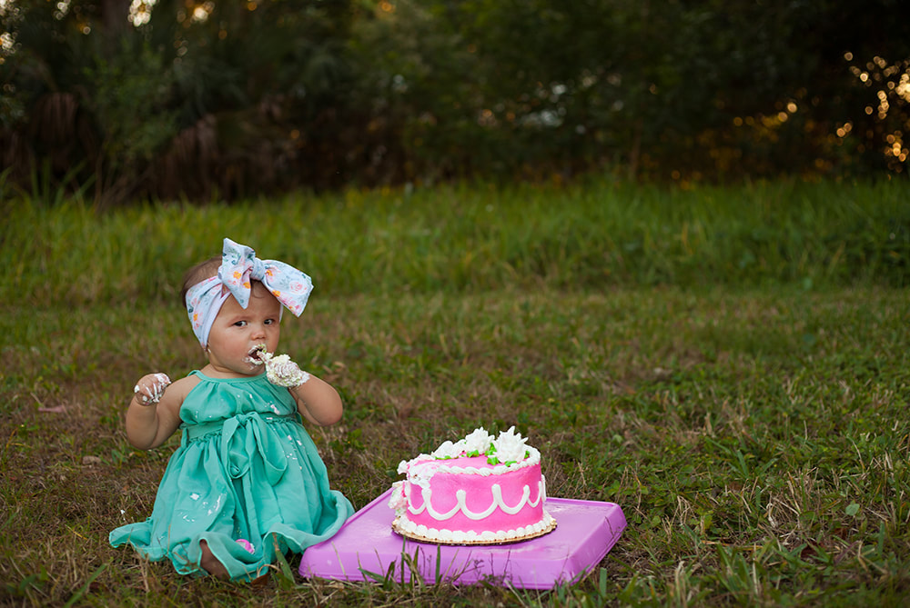 One year old baby girl in teal dress and big bow tastes her birthday cake with one finger