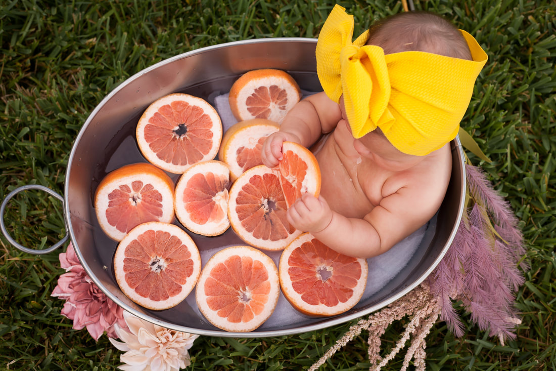 Shot from above, a baby with yellow bow sits in a tub of water filled with grapefruit slices