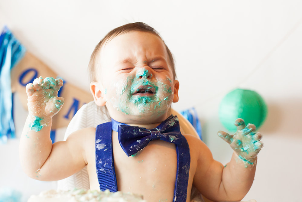 One year old baby boy in blue suspenders and bow tie with his face covered in blue cake frosting 