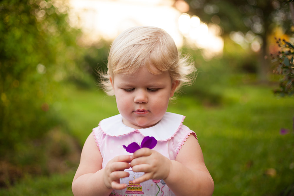 A toddler girl looks down at a flower she is holding in a dreamy environment 