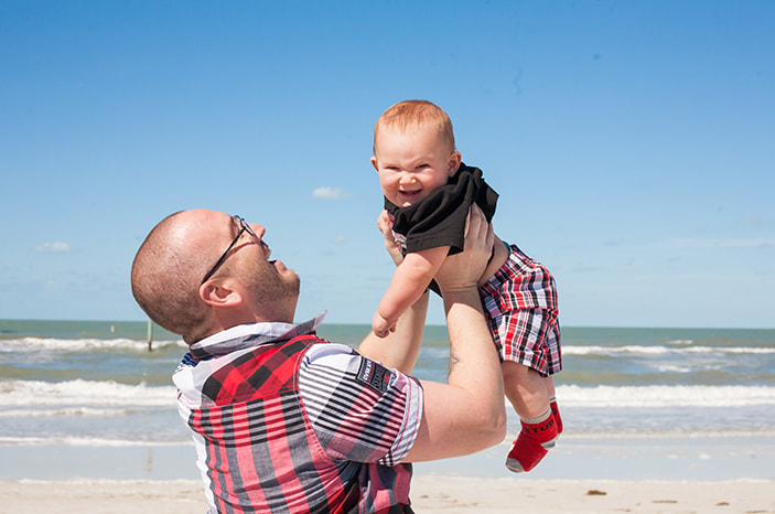 a father holds his baby in the air and smiles at him on a beach