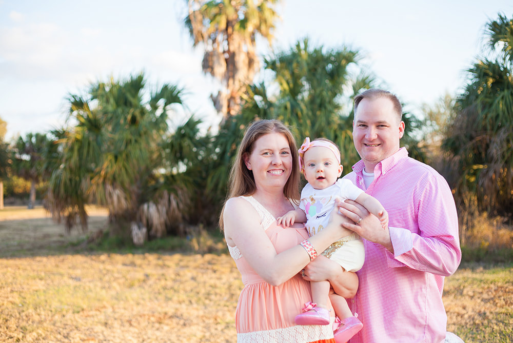Family of three smiles in the sun in front of a palm forest in Tampa Florida
