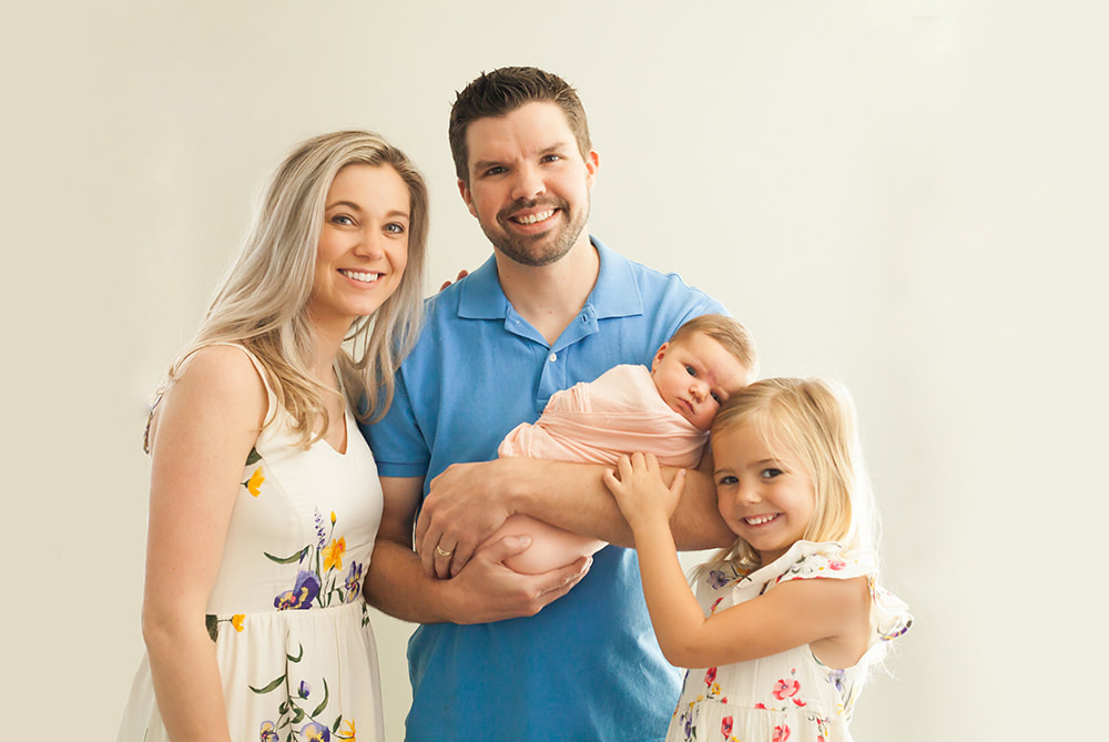 proud parents and big sister pose with newborn baby girl on off-white background