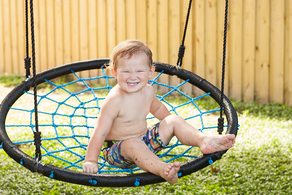 A toddler in swimming trunks smiles at the camera while swinging on a swing