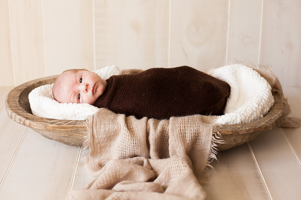 Newborn baby swaddled in brown lying in a long wooden bowl looks at the camera sweetly 