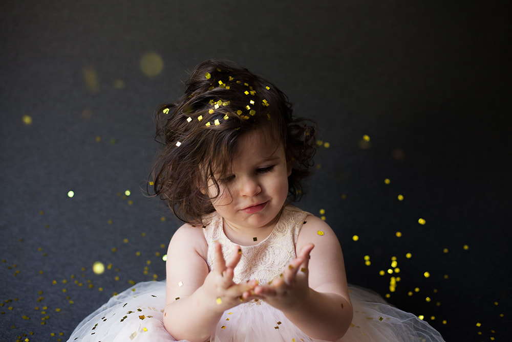 baby girl in pink tutu dress looks at her glitter covered hands while gold glitter falls all around her
