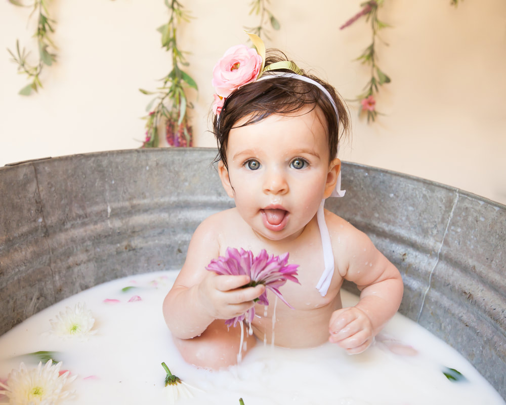 Close up of baby with bright eyes in a milk bath. She has her tongue out and is holding up a flower