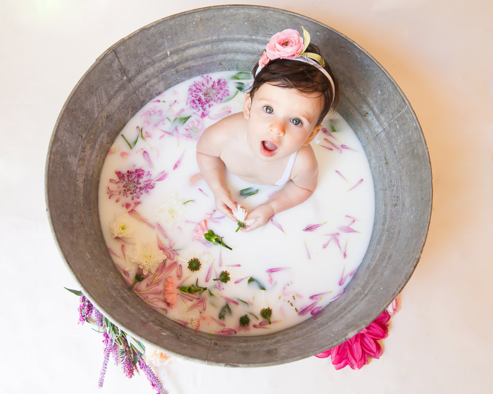 baby looks up at camera while sitting in a milk bath surrounded by flowers