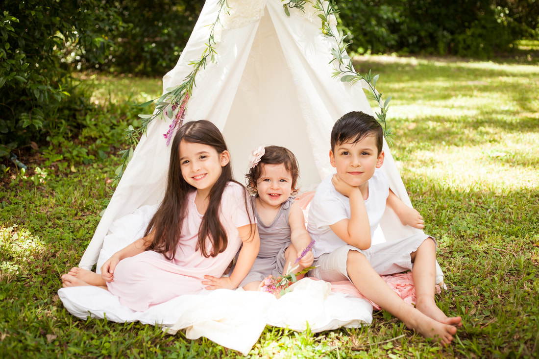 Three young children in light pink, white, and purple sit in the opening of a white tent draped in flowers