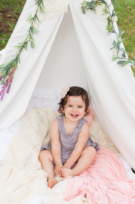 Smiling one year old baby sits in the opening of a white tent with flowers