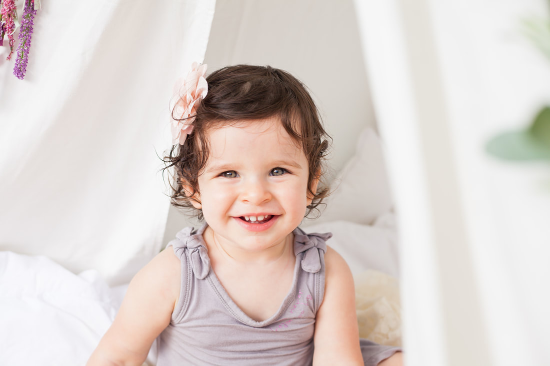 Close up of a smiling one year old baby in a purple dress surrounded by bright white draped fabric