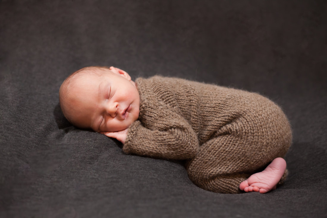 A sleeping newborn baby boy in brown knit pajamas lies curled up on his tummy