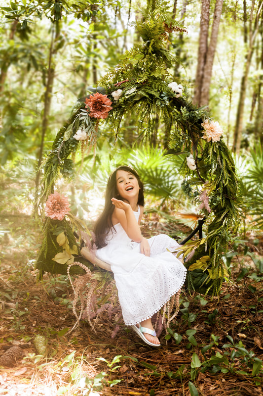a little girl in a flowing white dress smiles at the camera. She is sitting in a circle of flowers and foliage about 6 inches from the ground