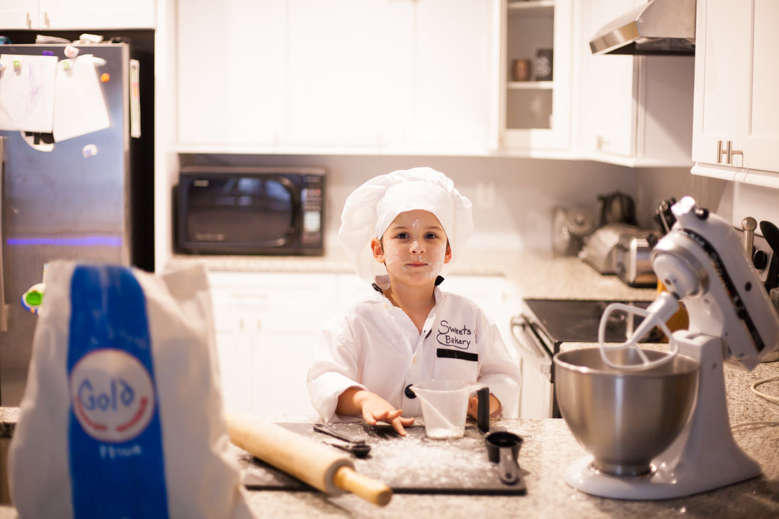 A little boy dressed as a baker is in a brightly lit white kitchen. He has flour on his face and baking tools on the counter around him