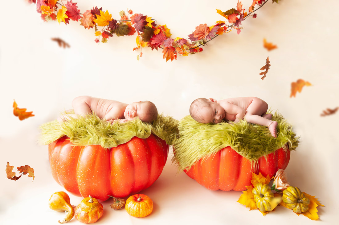 Twin newborn girls lie on two pumpkins with autumn leaves falling around them and a garland made of leaves behind them