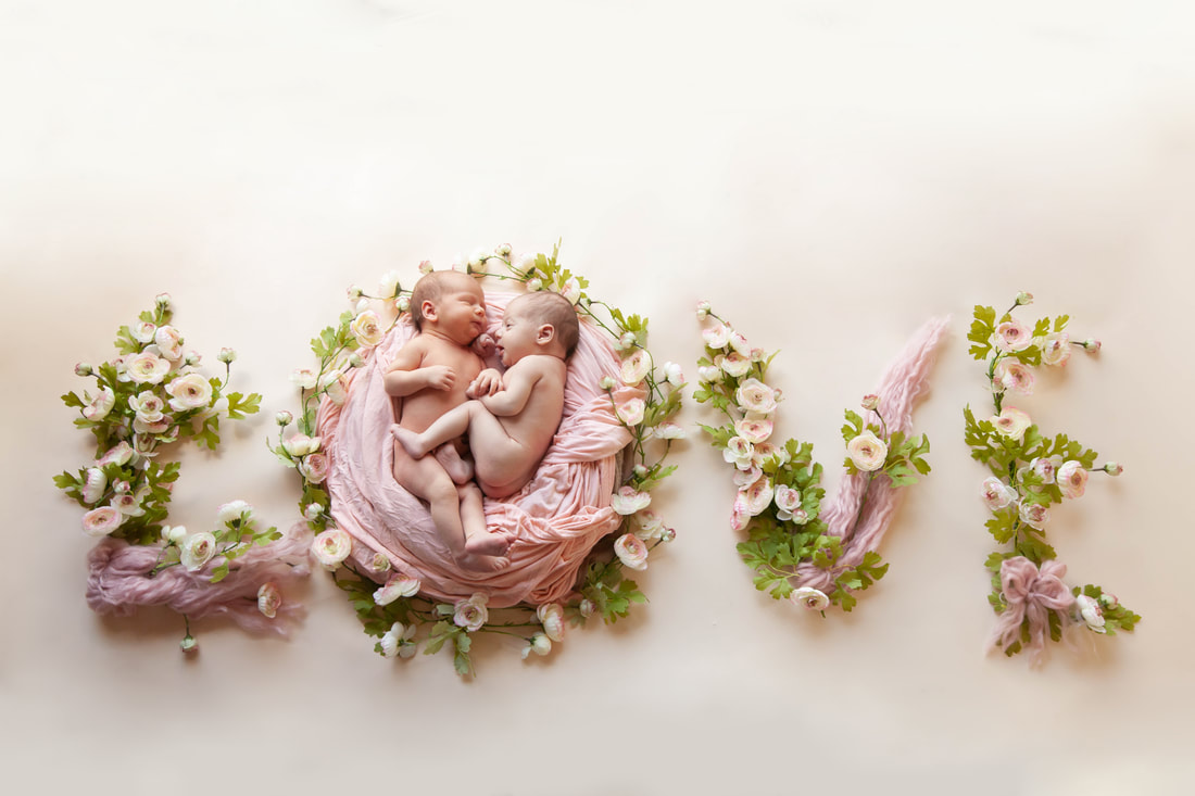 Newborn twins snuggle together in the O of the word love made out of pale pink flowers