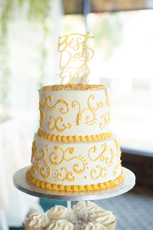 Photograph of wedding cake. White with yellow scroll work and a topper that reads 