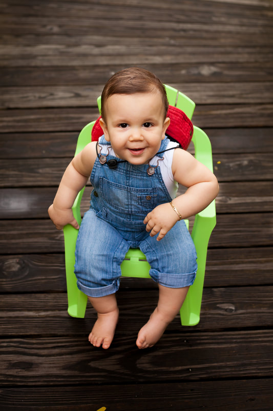 One year old boy in overalls sits in a bright green plastic chair on a dark wooden dock