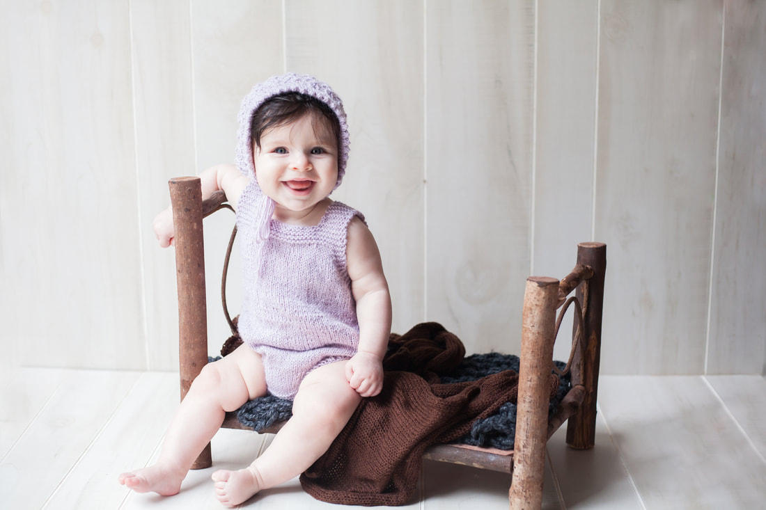 Six month old baby sits on bed in purple outfit and bonnet