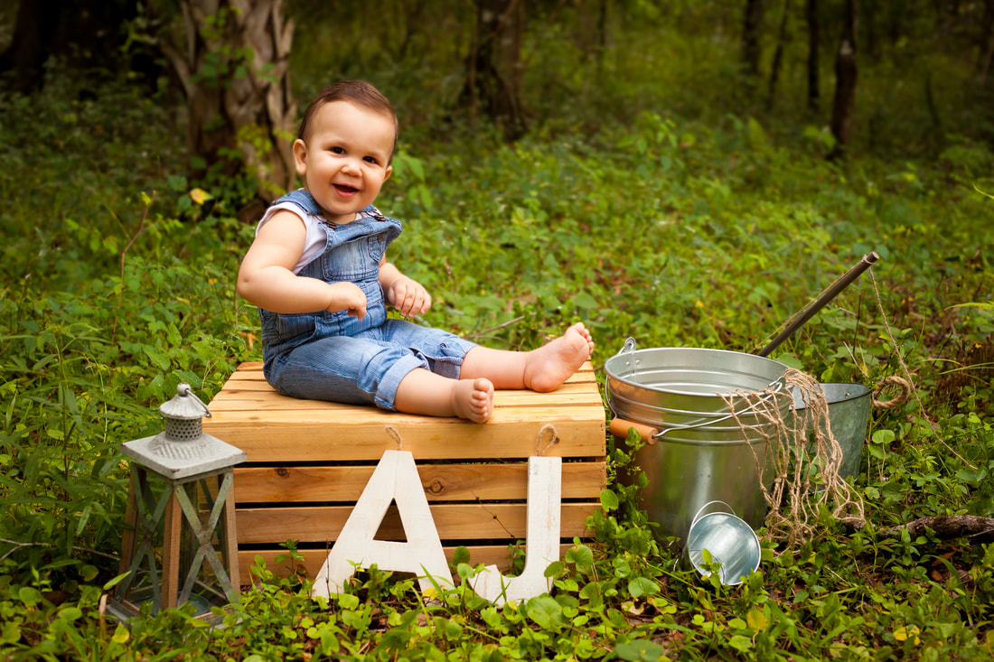One Year Old boy in overalls sits on a crate in lush greenery surrounded by fishing props