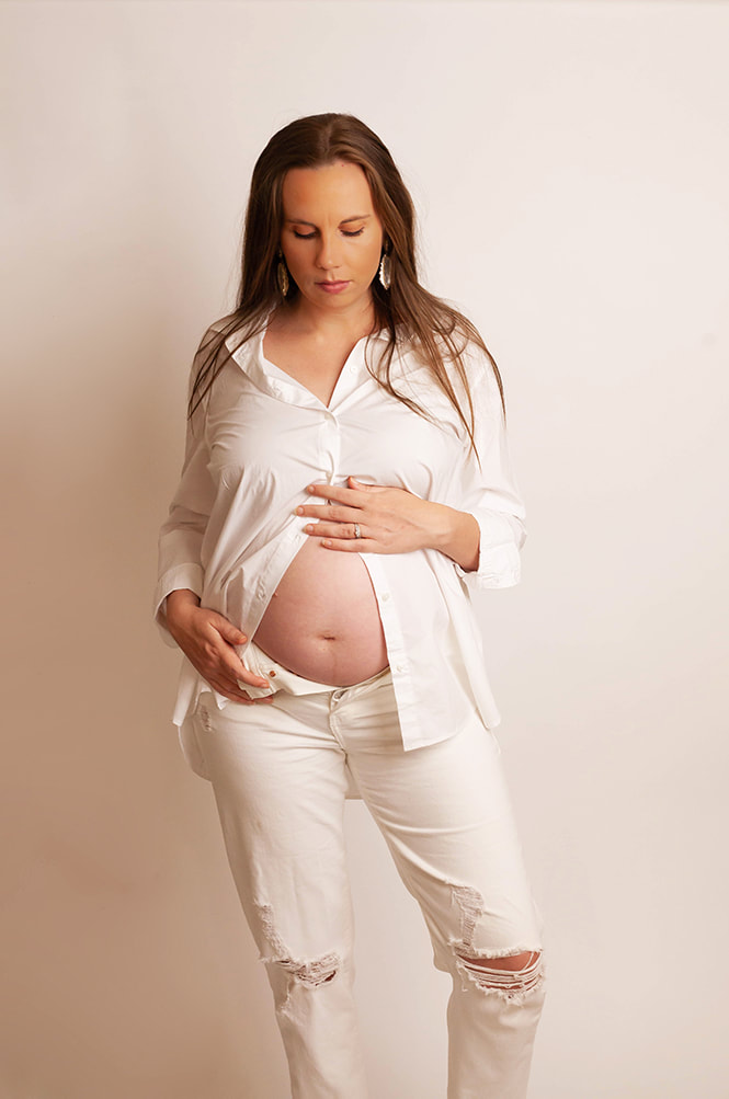 Pregnant woman looks down at her belly. She's on a white background and wears white jeans and a white button up shirt