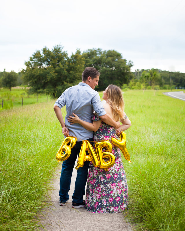 Couple holds balloons spelling BABY behind their backs in sunny field