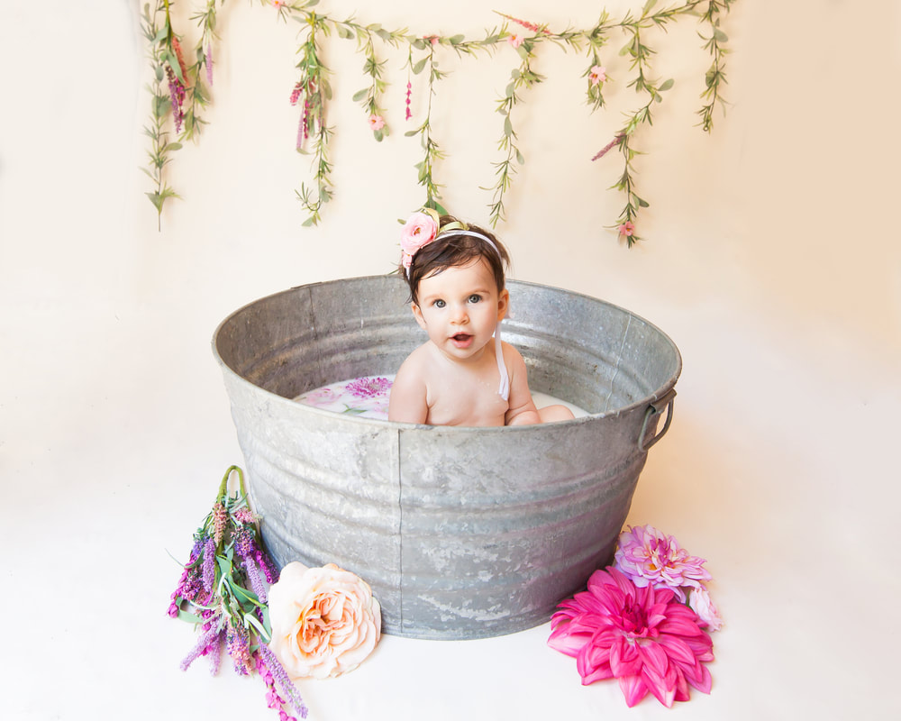 Baby girl looking in to the camera serenly surrounded by flowers while sitting in a galvanized tub filled with milk