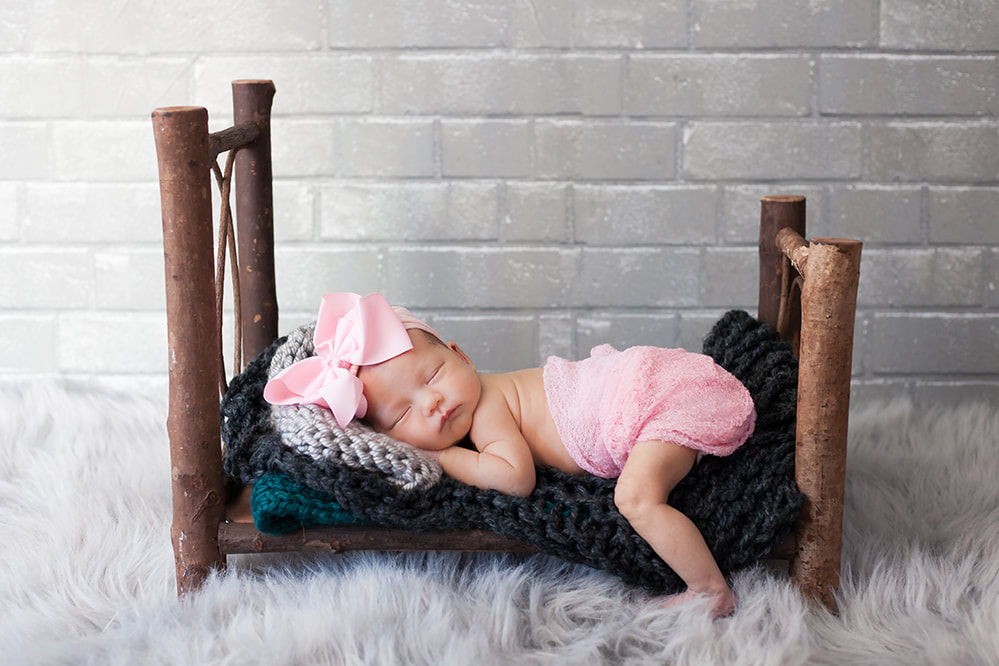Newborn baby girl with a big pink bow lies on a small wooden bed with a gray brick wall behind her