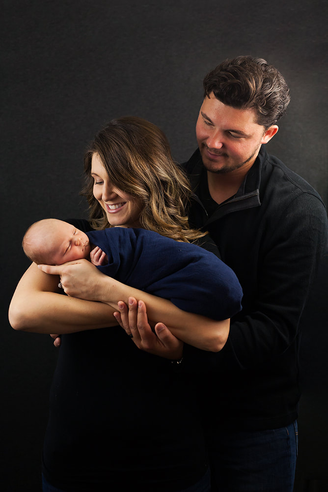 Two parents smile at their newborn baby in front of dark gray backdrop