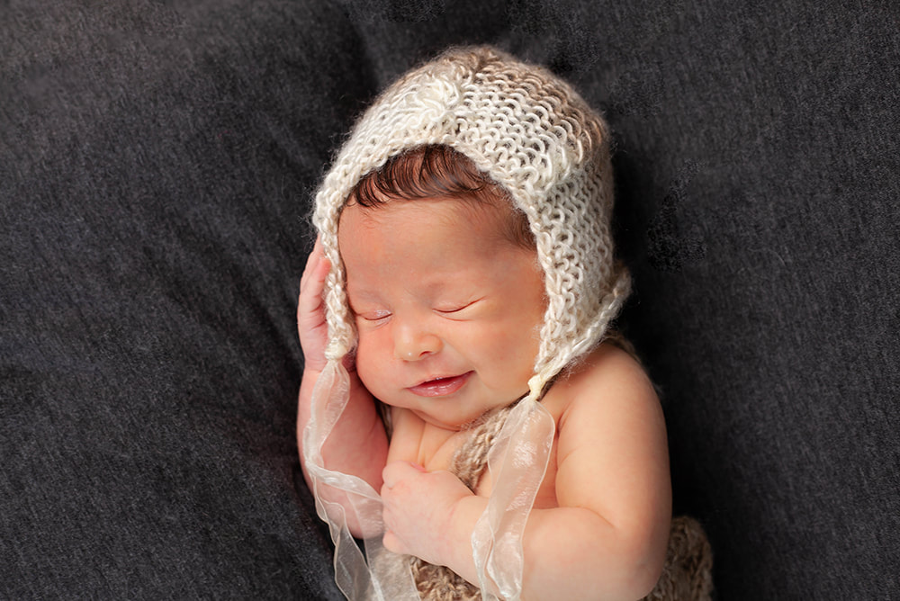 A newborn baby girl smiling with eyes closed wearing a tan and brown bonnet
