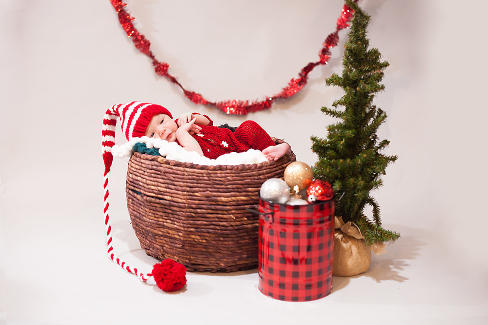 Newborn baby boy in red pajamas lies in a brown woven basket wearing a long tailed elf hat with red and white stripes. a tiny Christmas tree stands next to the basket and a red and black tin bucket full of Christmas ornaments is next to it. behind him hangs a red garland on a white background 