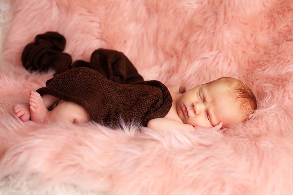 Newborn baby girl sleeping on Pink fur wrapped in a brown knit swaddle
