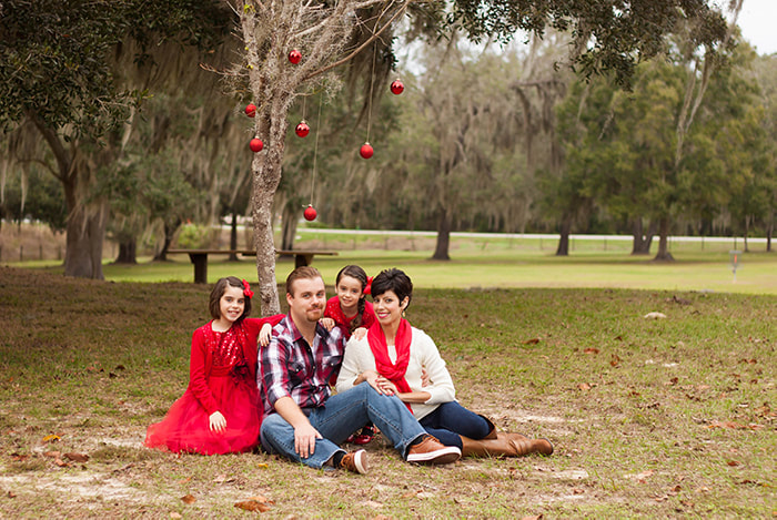 family of four sit at the foot of a tree with red christmas bulbs hanging over them
