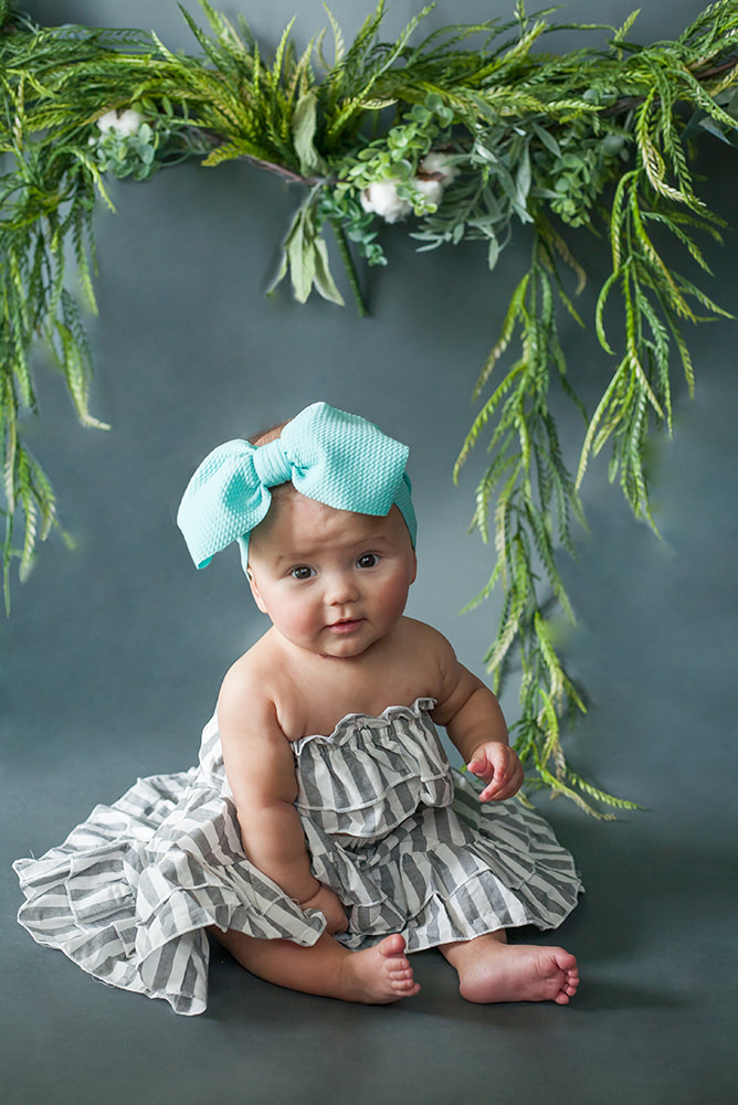 baby girl on a dark gray background with foliage all around her in gray and white dress