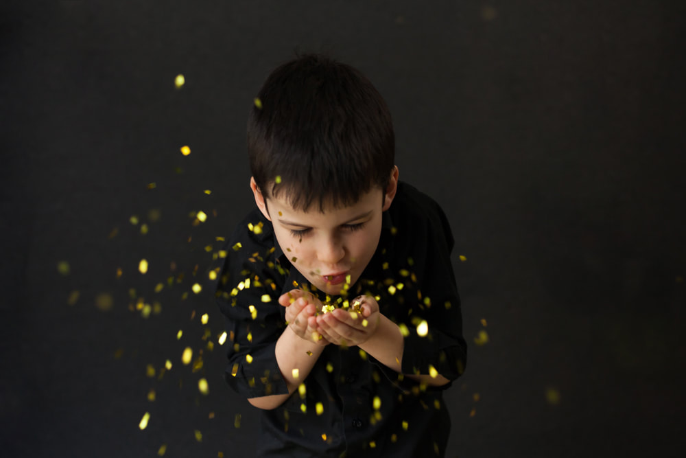 Little boy on a black background Blows bright gold glitter out of his cupped hands toward the camera