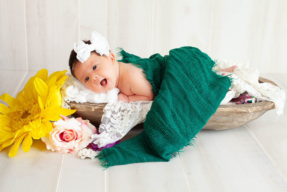 A newborn baby girl in a long wooden bowl looks at the camera while surrounded by brightly colored flowers