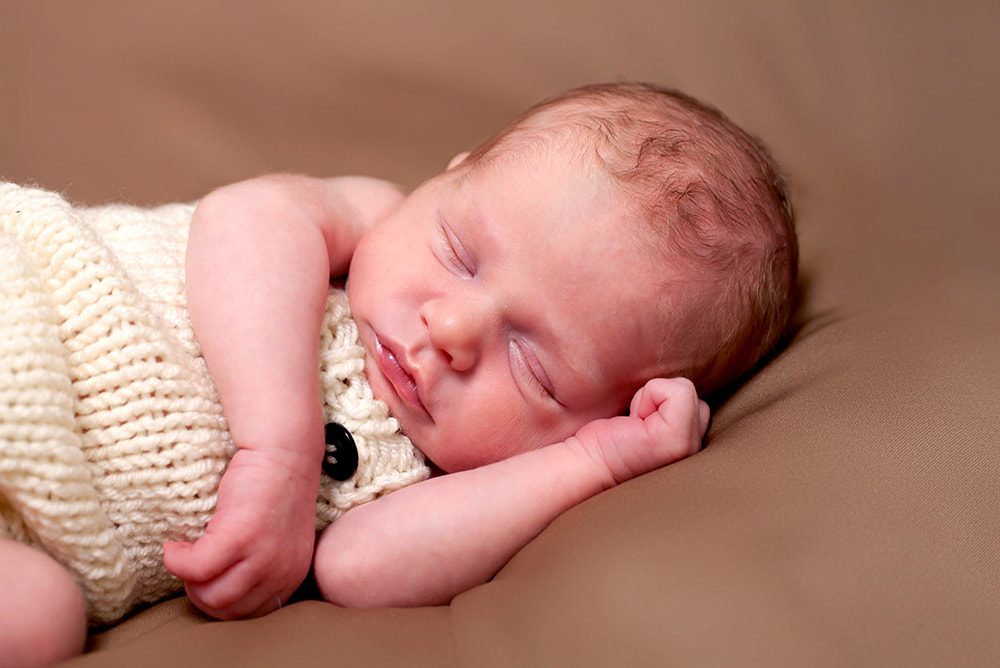 Newborn baby boy wearing white knit overalls sleeps on his side on a light brown backdrop