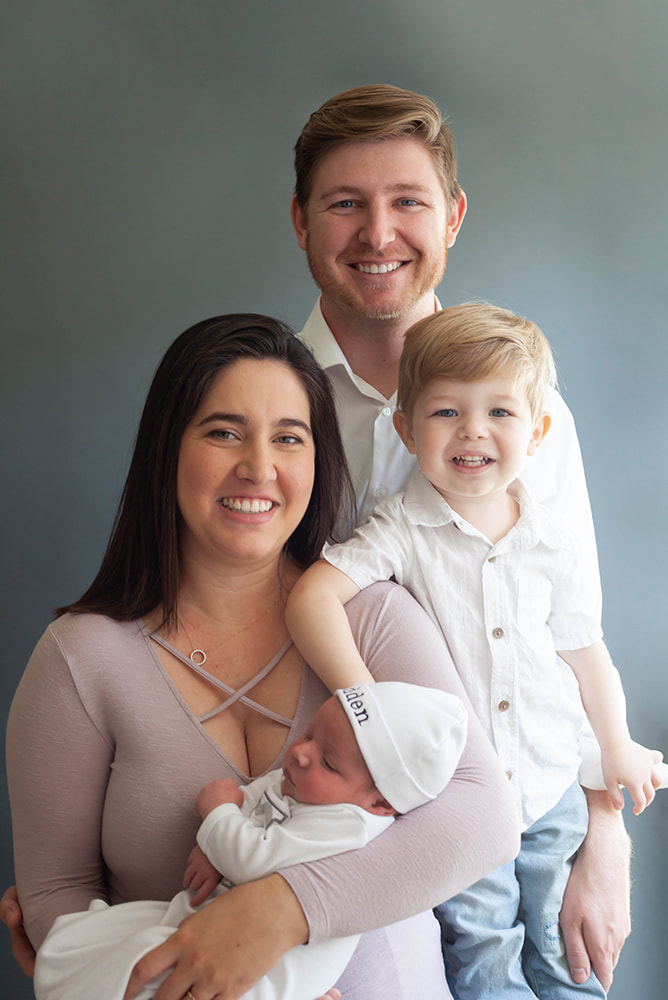 Parents pose with their newborn baby and toddler on a slate Gray background