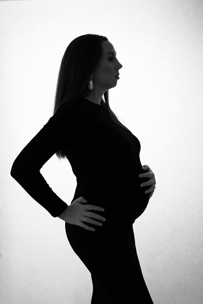 Pregnant mother with one hand on her belly and one on her hip in profile. She wears a black dress and looks straight ahead