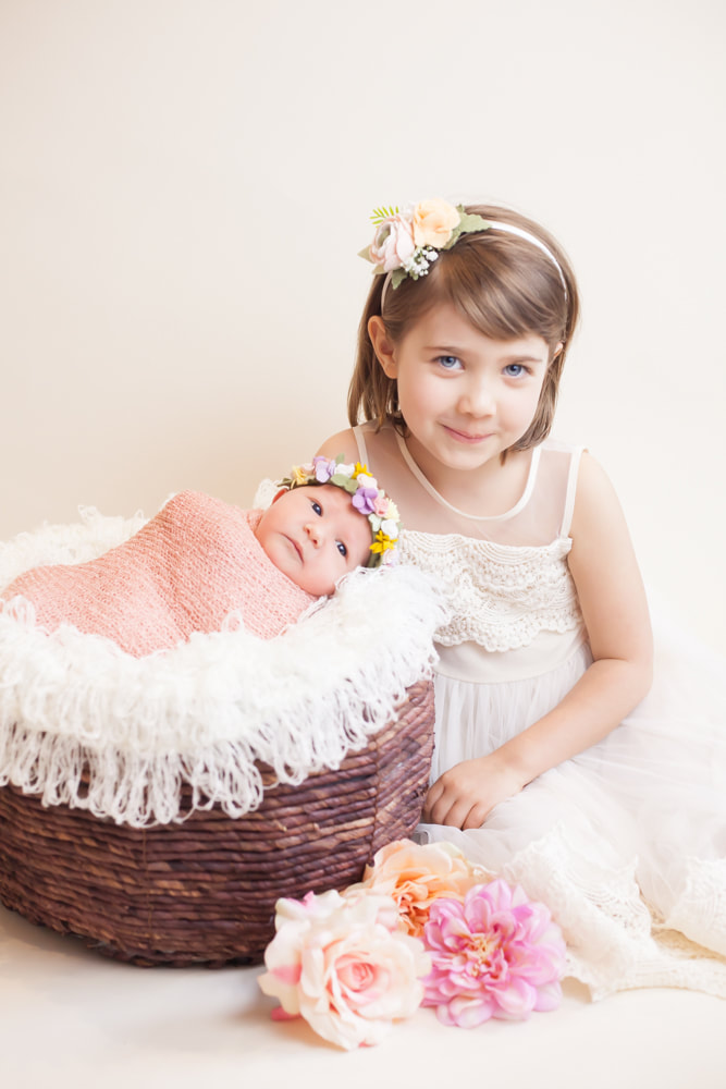Big sister smiles next to newborn baby in a basket on a white background. Taken in Tampa, FL