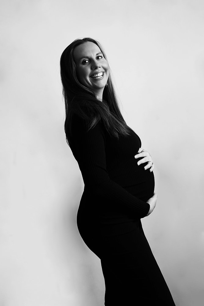 Pregnant woman in a black dress smiles exhuberantly over her shoulder while holding her belly