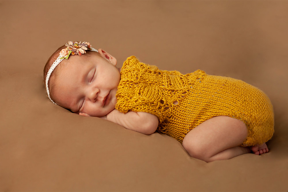 Newborn baby in yellow with a flowered headband sleeps on a brown background