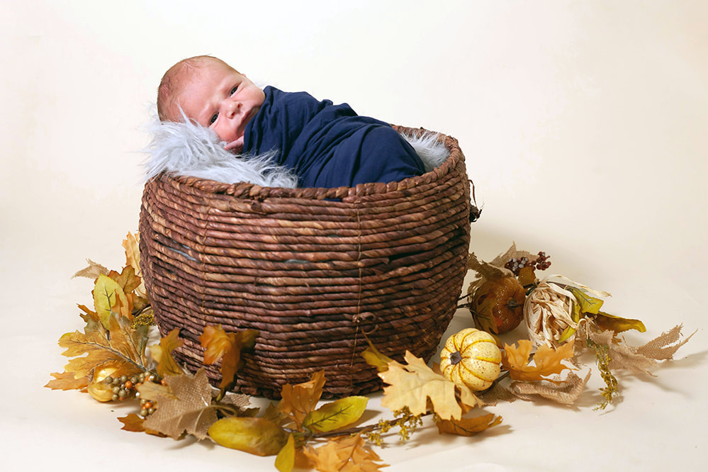 baby boy lying in a brown woven basket surrounded by fall leaves and pumpkins
