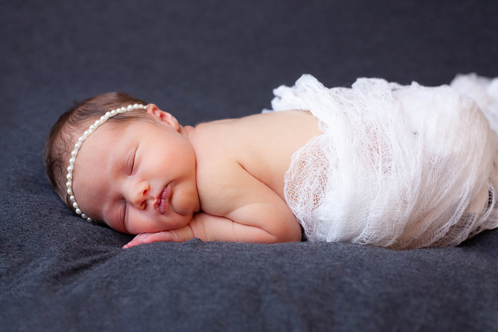 Newborn baby wearing a pearl headband and wrapped in white swaddle on a dark gray background