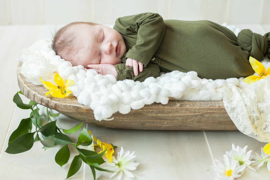 baby in green sleeps on her side in a wooden bowl surrounded by flowers and greenery